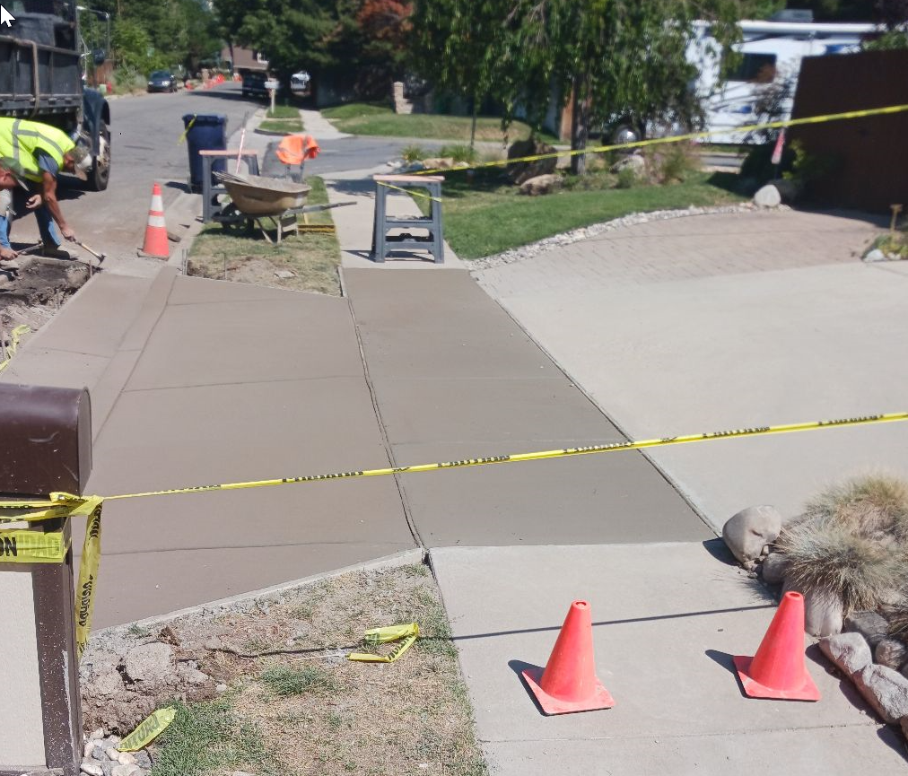 Sidewalk construction zone with cones, caution tape, and workers repairing a cracked path.