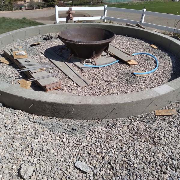 Outdoor fire pit in a gravel area, with a large metal bowl and blue hose on wooden planks.