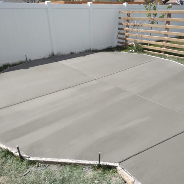 Empty concrete backyard patio beside a white fence and wooden privacy screen, with a small grassy edge