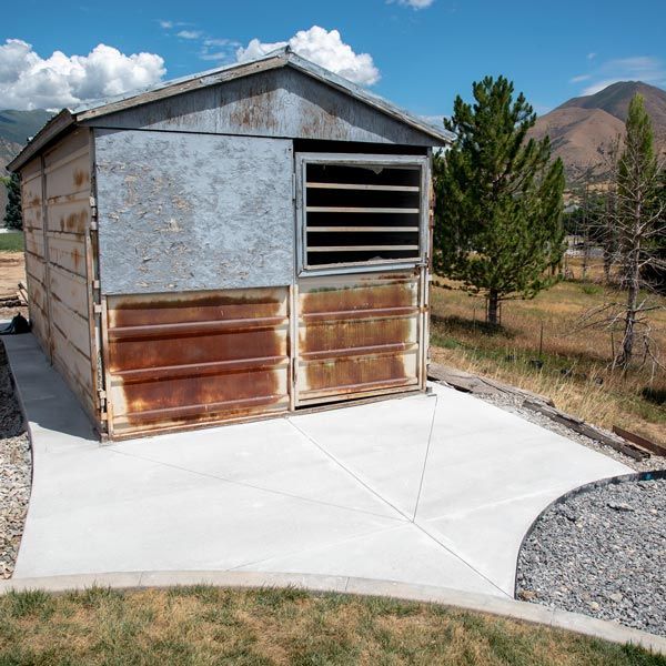 Small metal shed with a rusted front, set on a concrete pad in a rural yard with mountains in the background