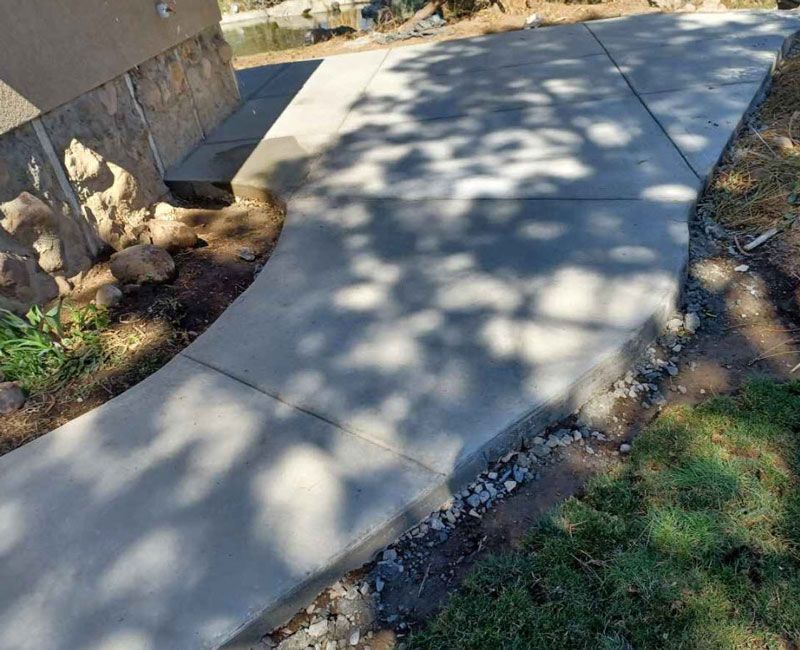 New concrete sidewalk curves beside a stone wall, with tree shadows across the path.