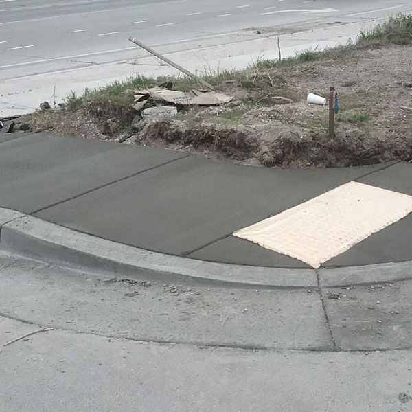 Sidewalk curb ramp with a beige tactile panel beside a street and dirt verge.