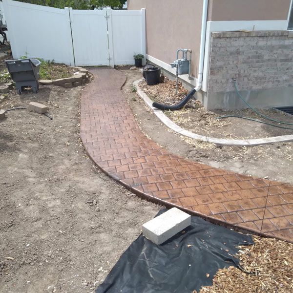 Curved brick walkway beside a house under construction, with bare dirt and a white fence in the background