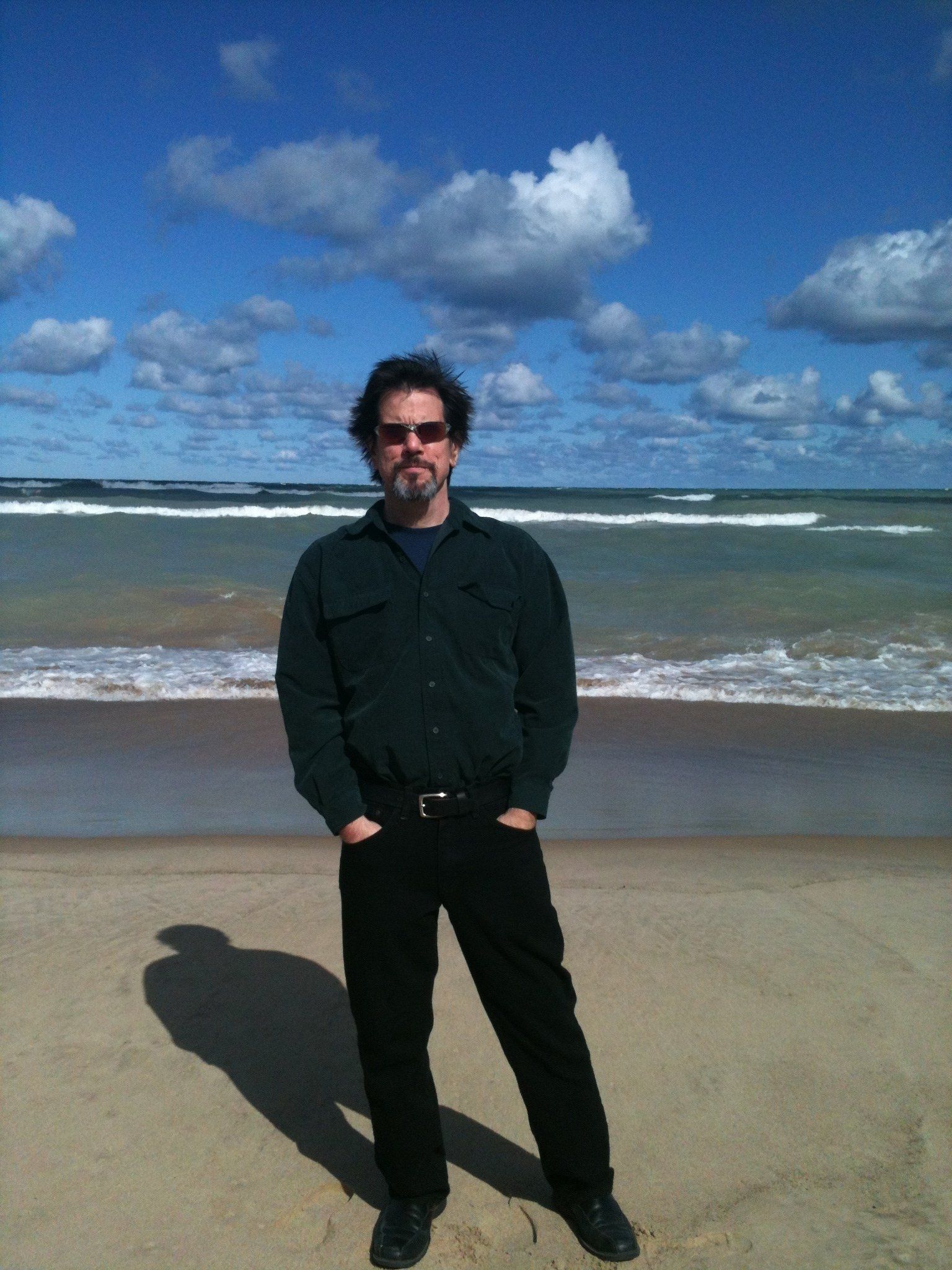 Person standing on a sandy beach under a cloudy blue sky, wearing dark clothes and sunglasses.