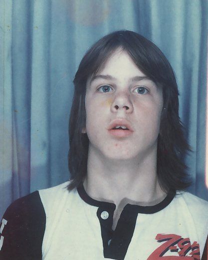 Person with shoulder-length dark hair in a white-and-black shirt, standing against a blue curtain, looking at camera.