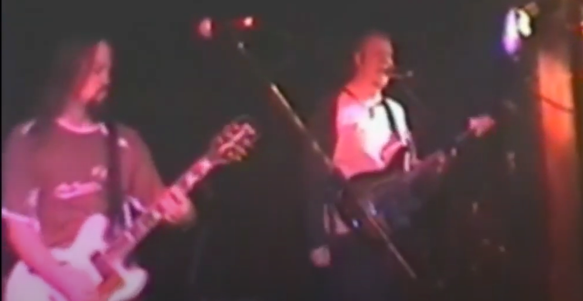 Two musicians performing on a dimly lit stage with guitars under pink and red lights