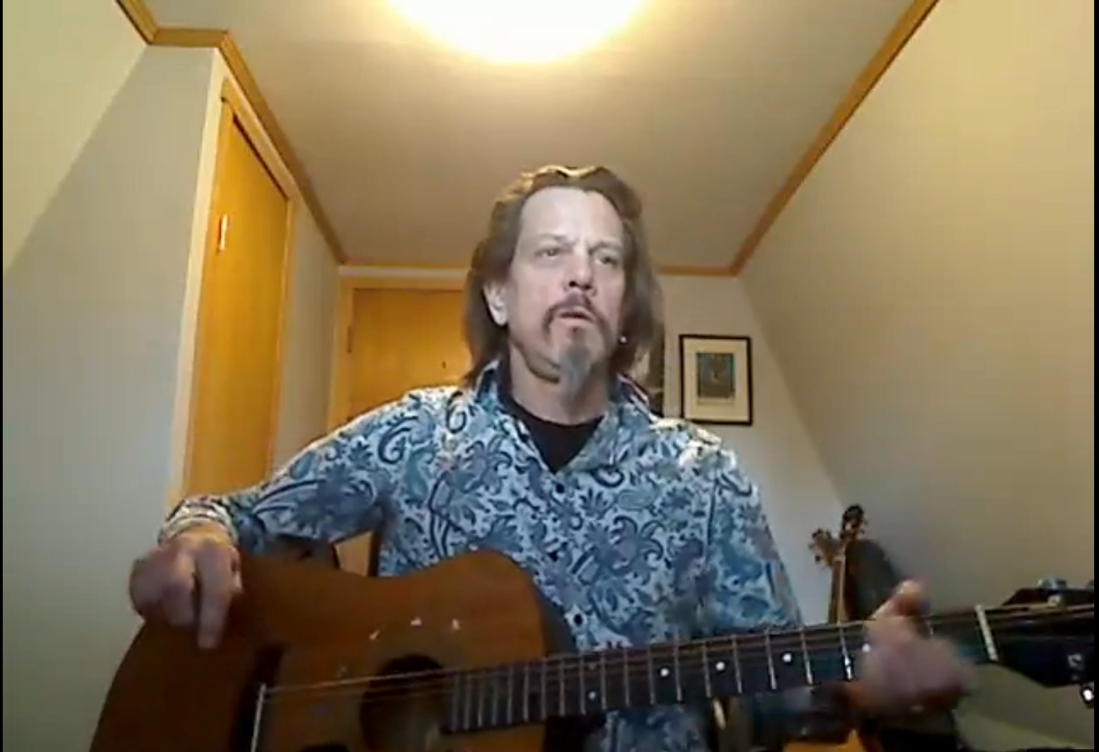 Man playing acoustic guitar in a small room with warm lighting and a framed picture on the wall