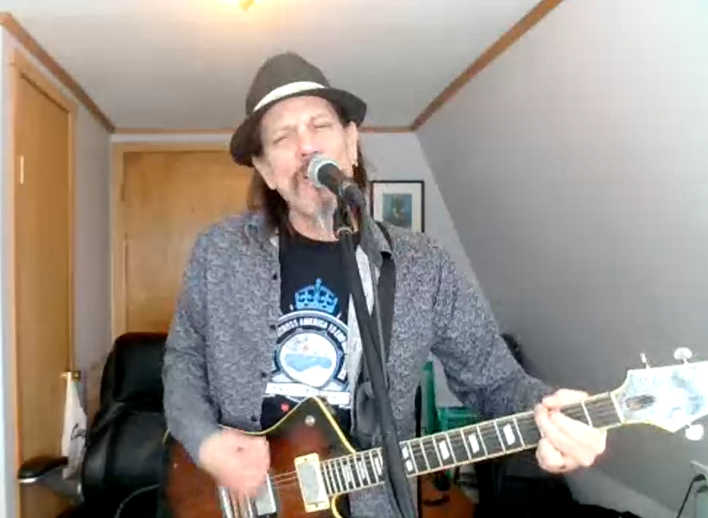 Man singing into a microphone while playing electric guitar in a home room with slanted ceiling