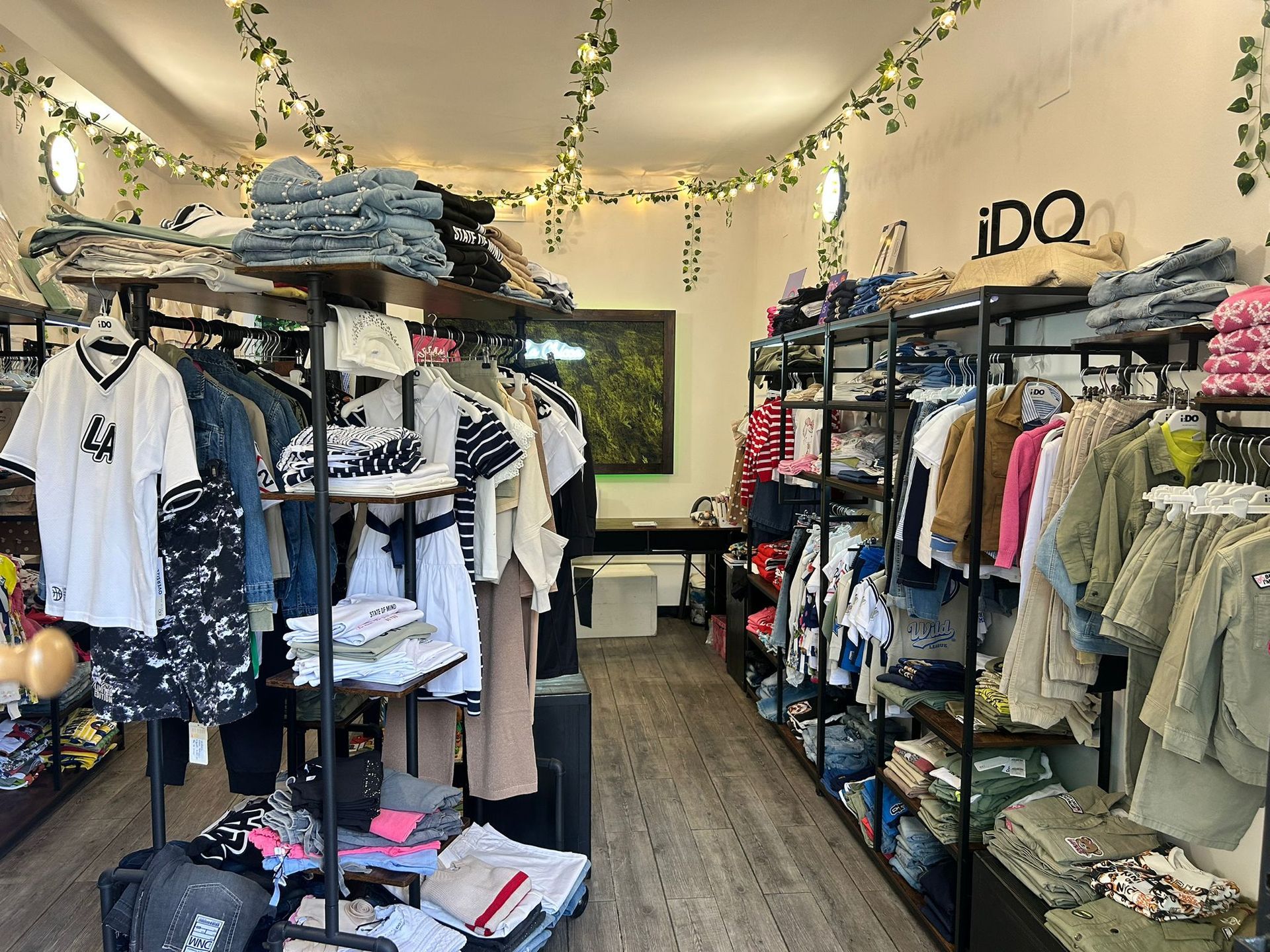 Interno di una boutique di abbigliamento con scaffali pieni di vestiti piegati e appesi, illuminati da luci calde e con piante rampicanti.