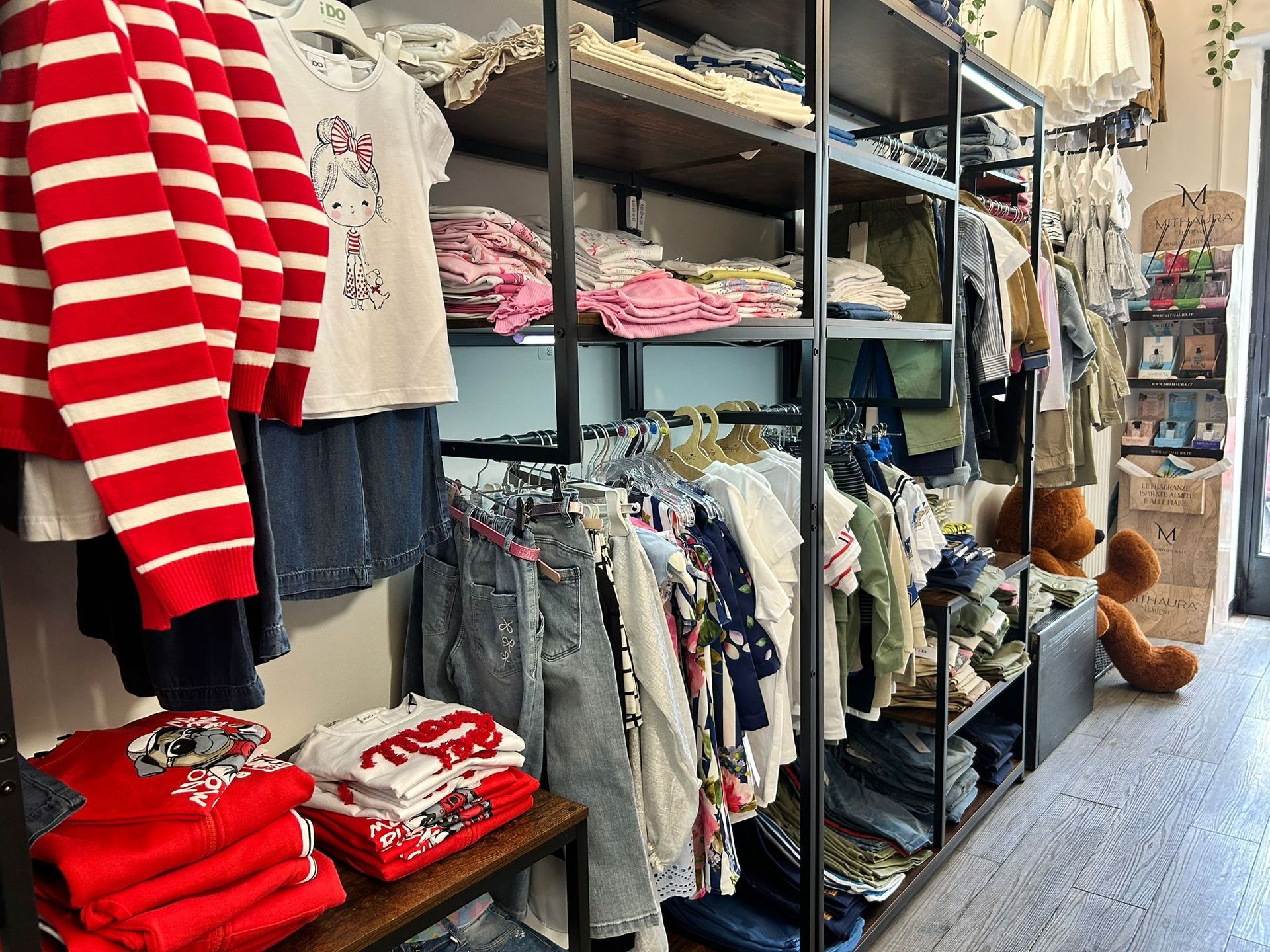 Corridoio di un negozio di abbigliamento con maglioni piegati, camicie appese e orsacchiotti di peluche sugli scaffali lungo uno stretto passaggio