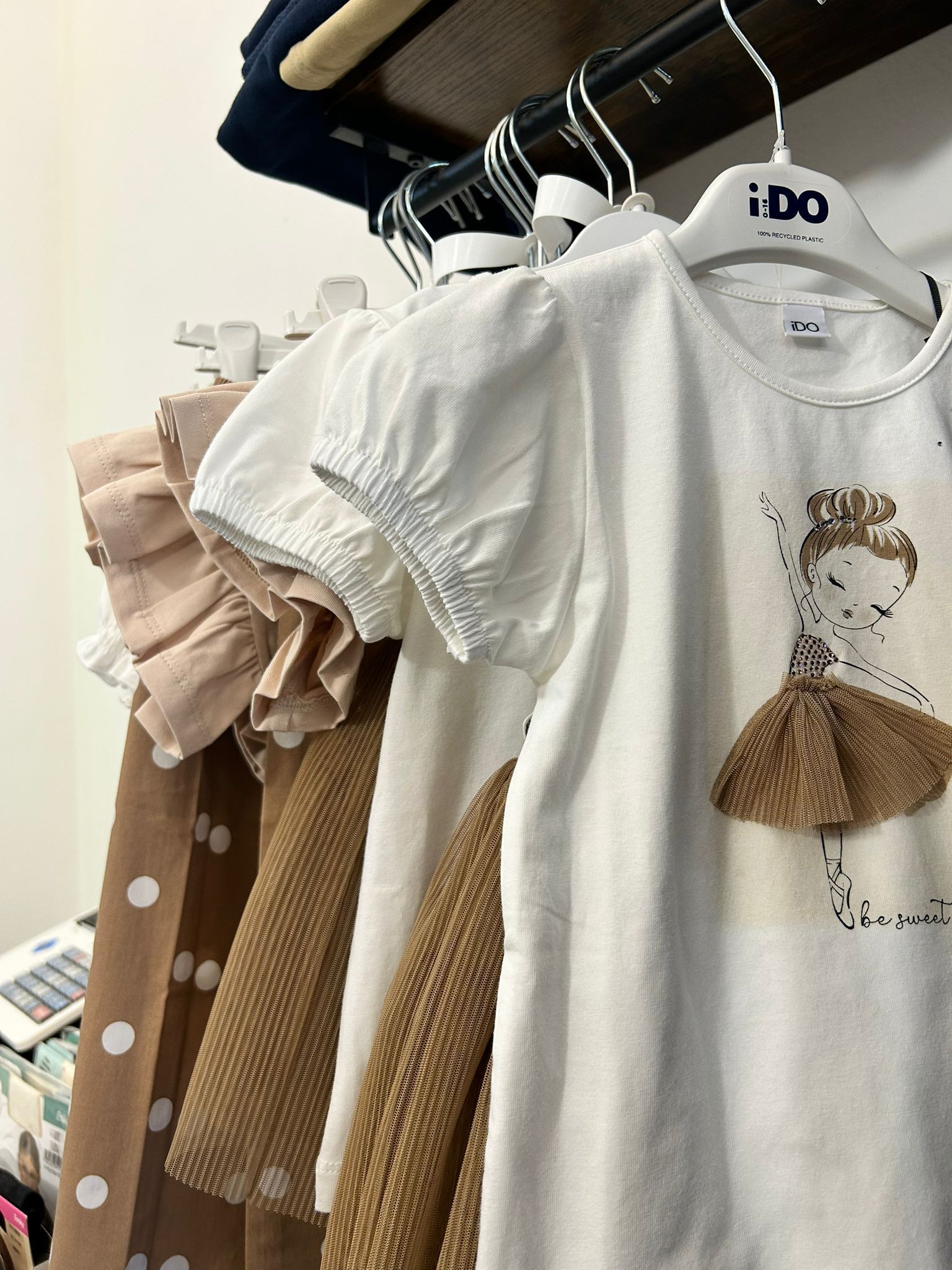 Vestiti per bambini appesi a uno stendino, tra cui una maglietta bianca con stampa e un vestito beige con volant.