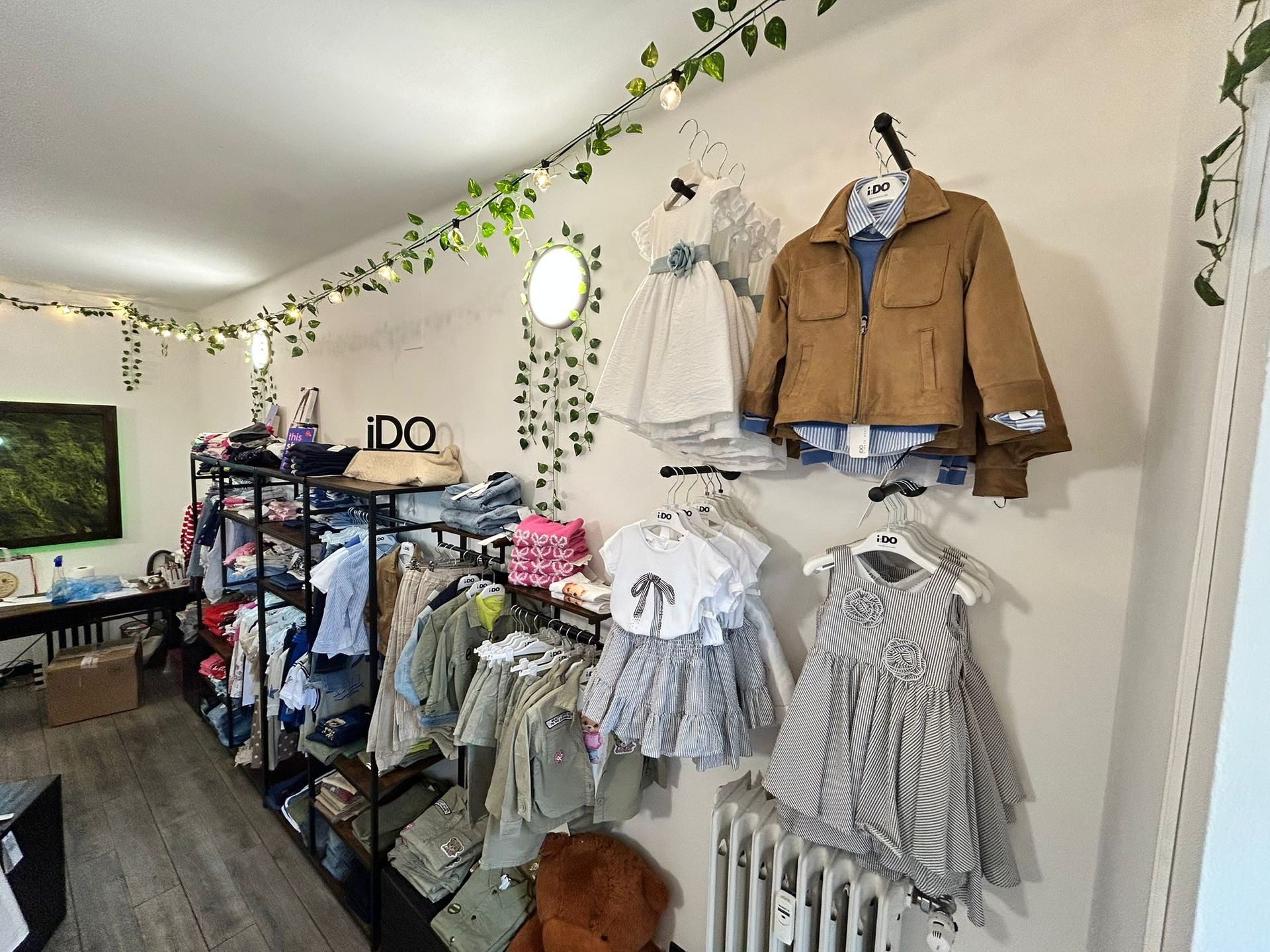 Interno di un negozio di abbigliamento con scaffali pieni di vestiti per bambini, un espositore di giacche color cammello e ghirlande di rampicanti lungo la parete.