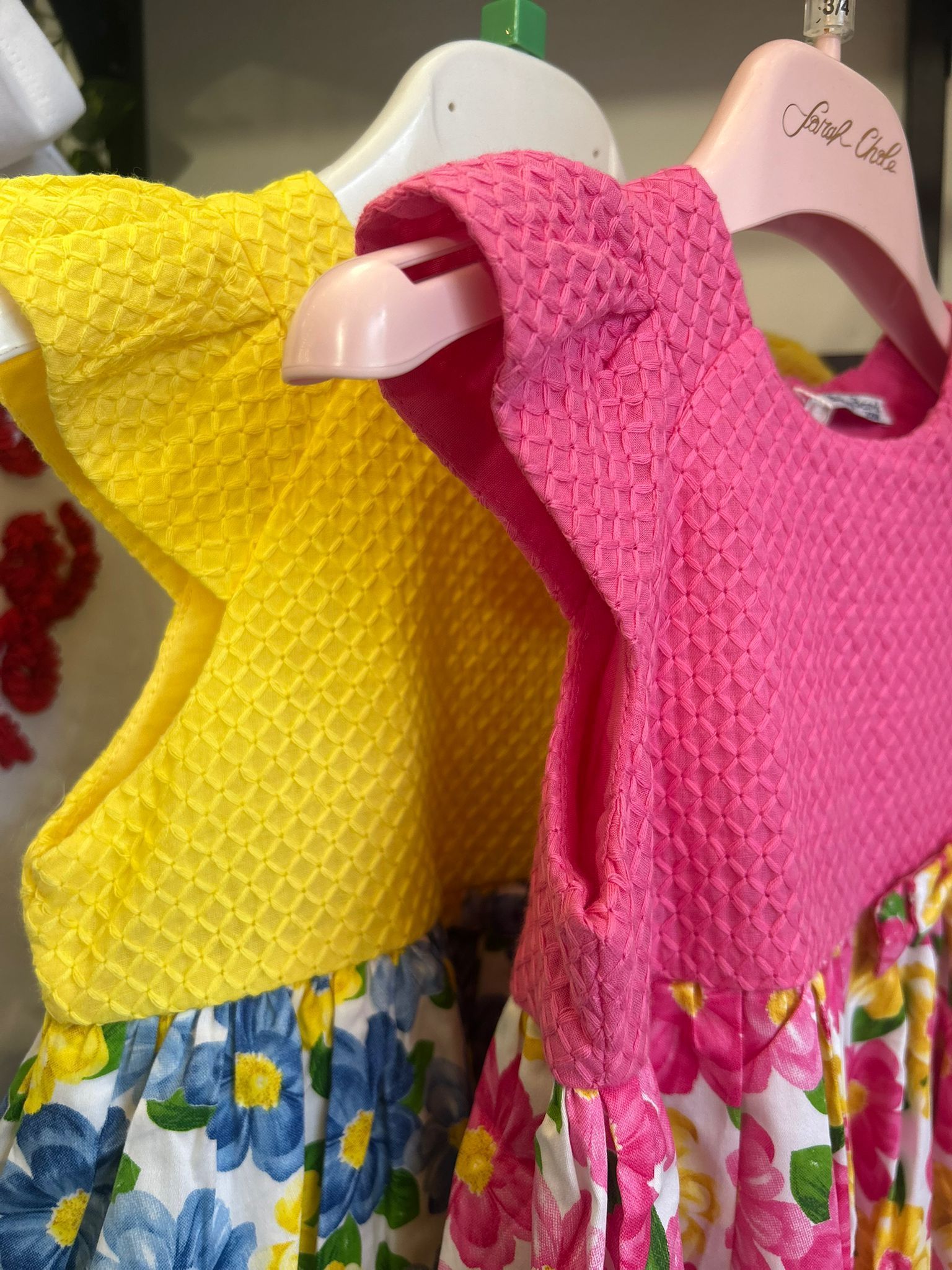 Due coloratissimi vestitini da bambina, uno giallo e uno rosa con gonne a fiori, sono appesi uno accanto all'altro su delle grucce.