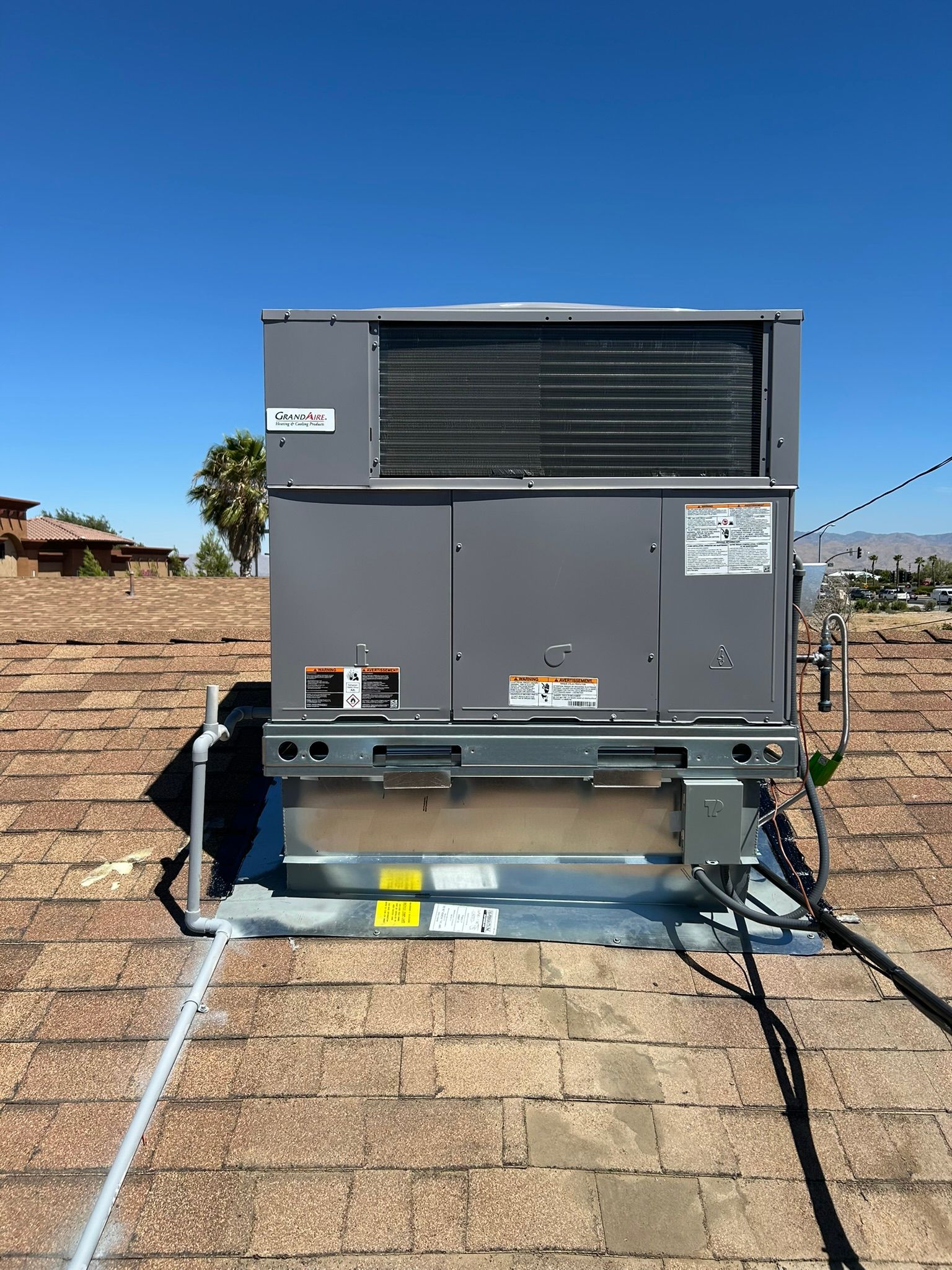 Rooftop air conditioning unit with a gray exterior on a brown shingle roof under a blue sky.
