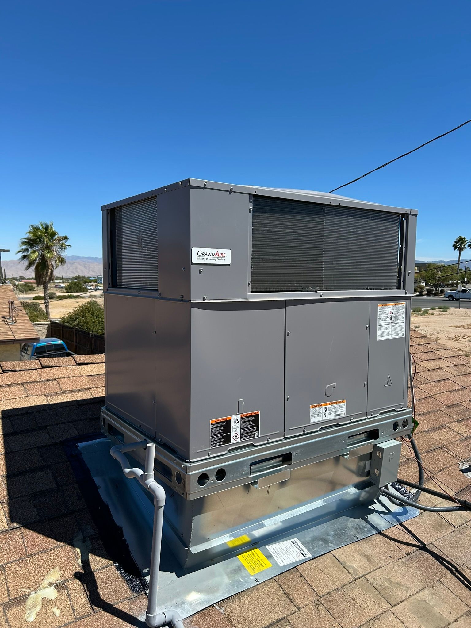 Rooftop HVAC unit on a sunny day. Gray metal box, dark grilles, mounted on a silver base, with clear blue sky.