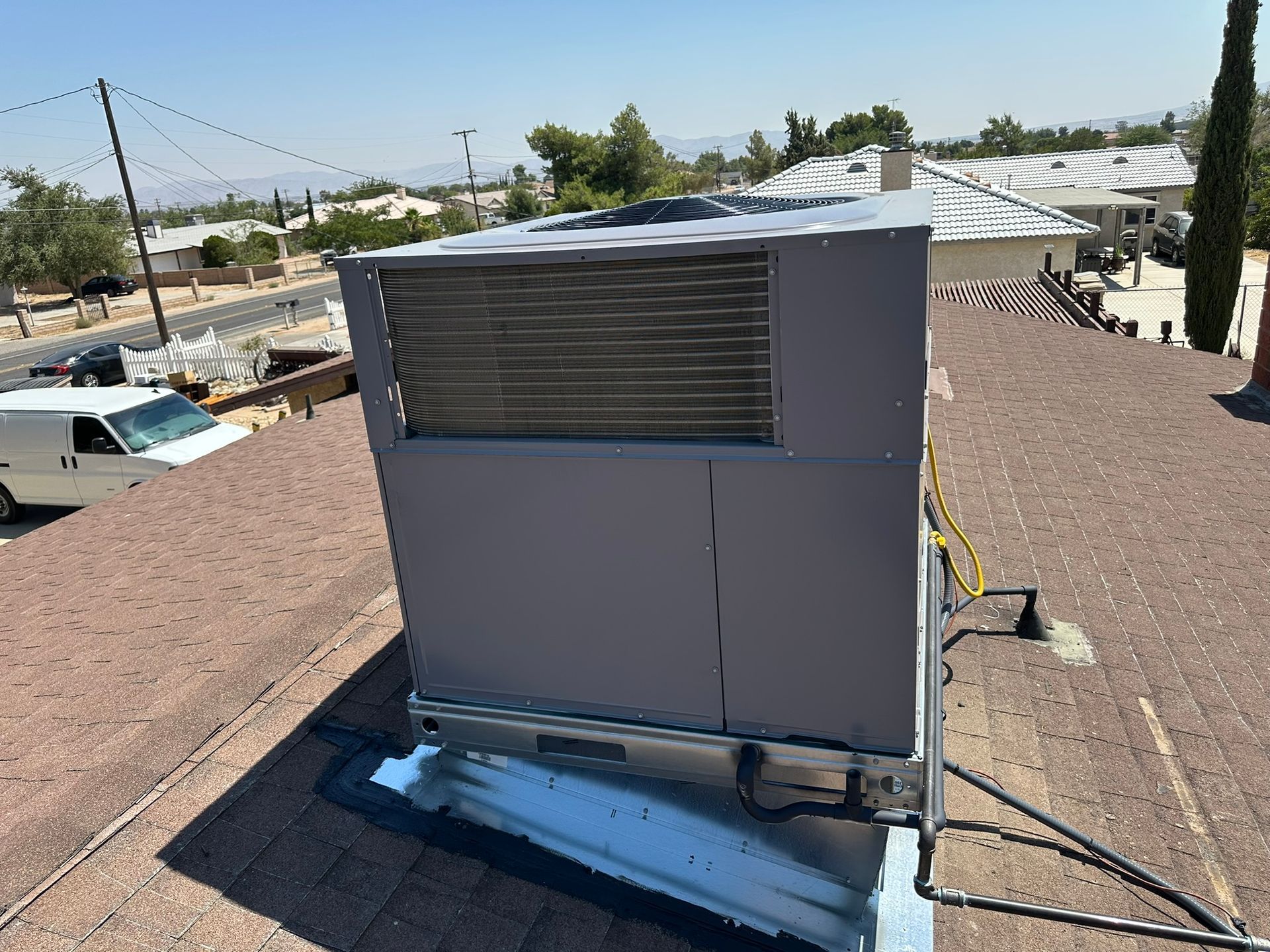 HVAC unit on a rooftop. Grey metal unit with fins, brown roof, white van, and neighborhood in the background.