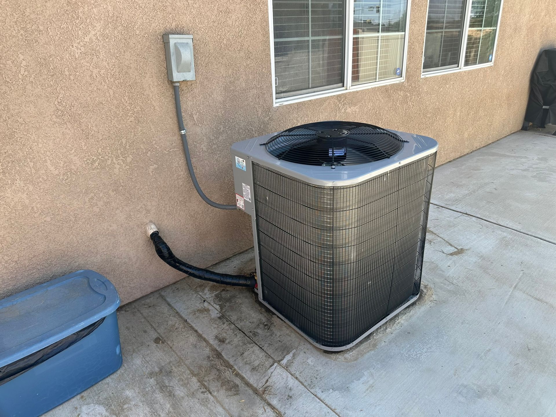 An air conditioning unit next to a stucco wall and a storage container on a concrete surface.