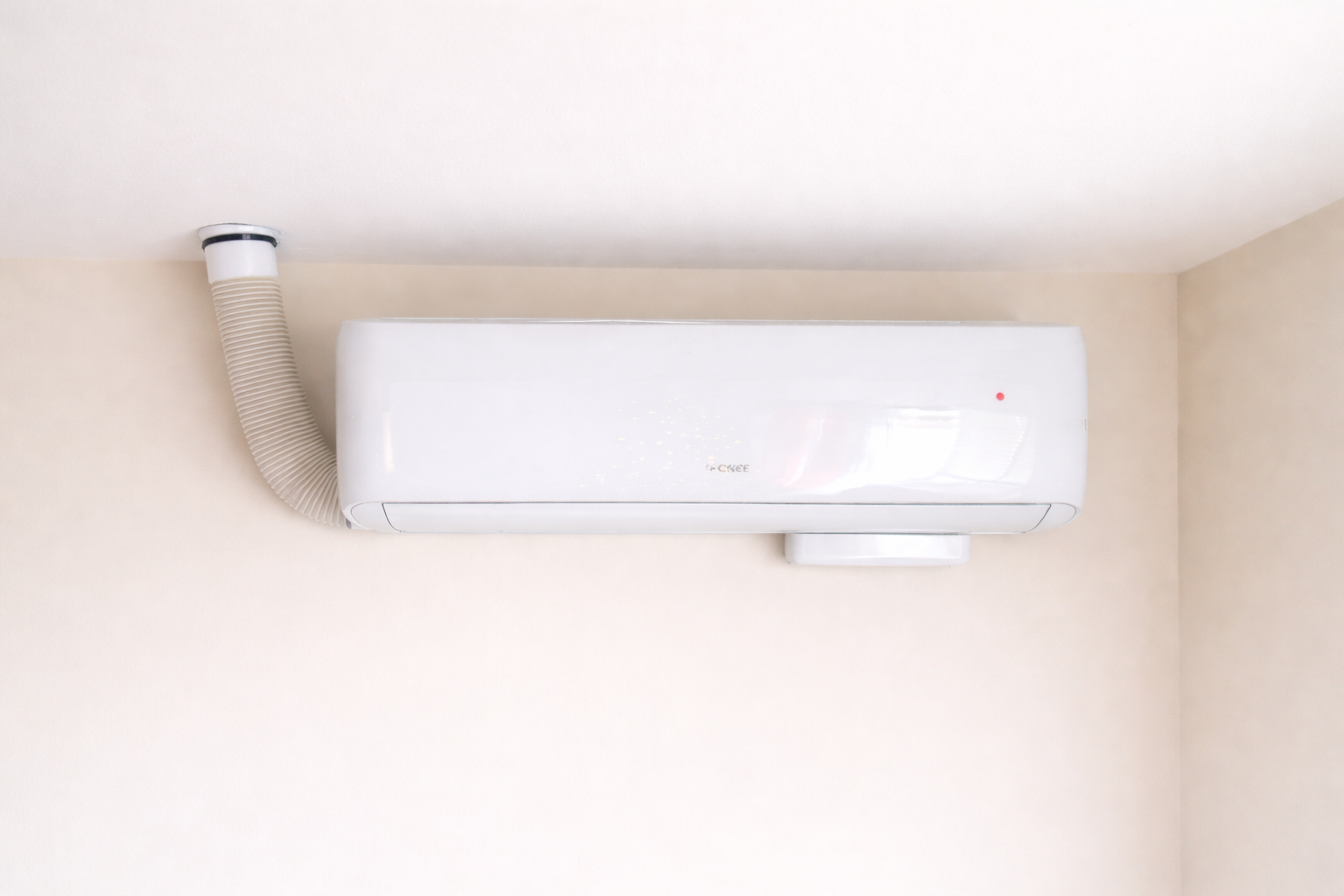 White air conditioner unit mounted on a beige wall, with a flexible exhaust tube connected.