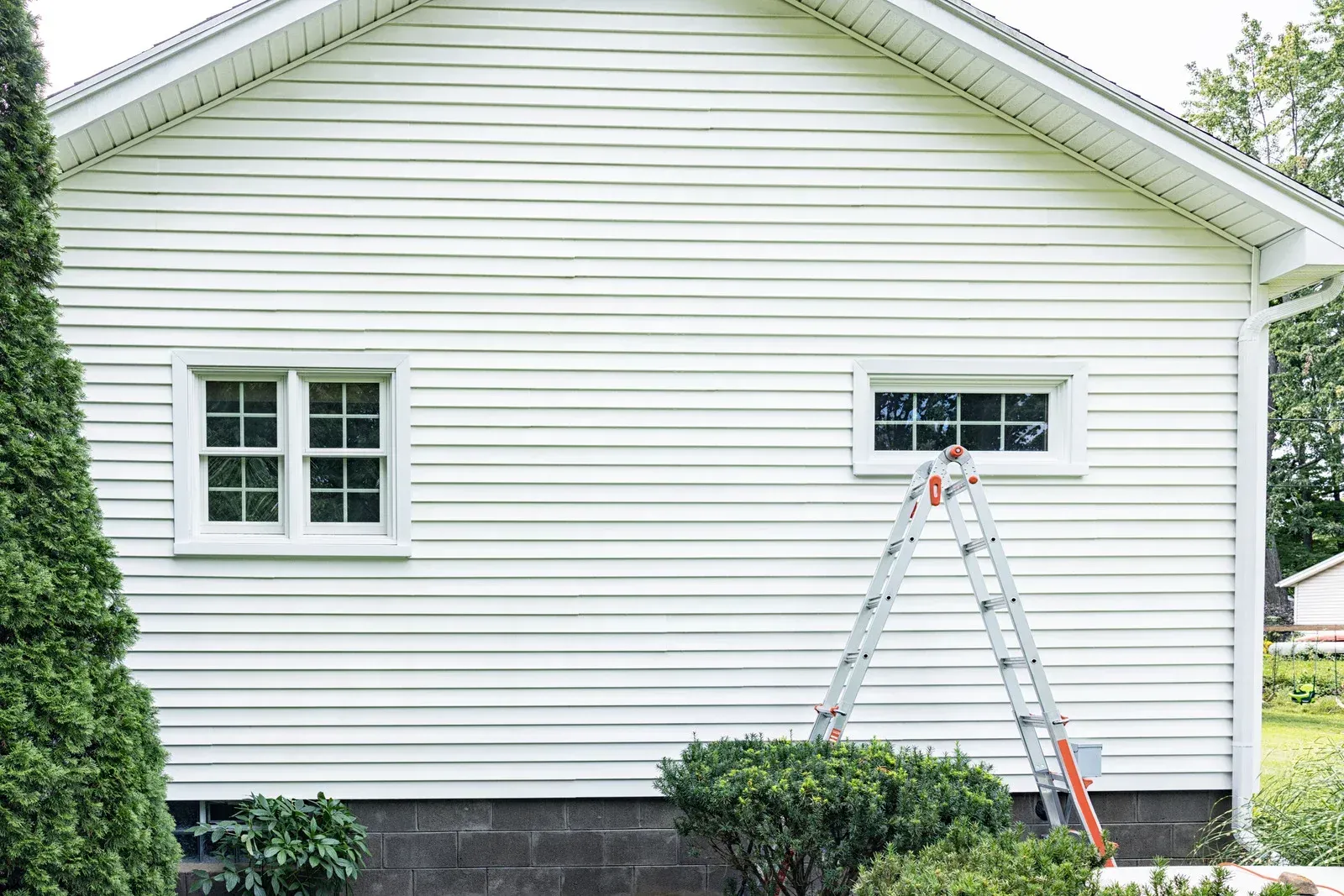 White house siding with two windows; a ladder leans against the side near one window, greenery below.