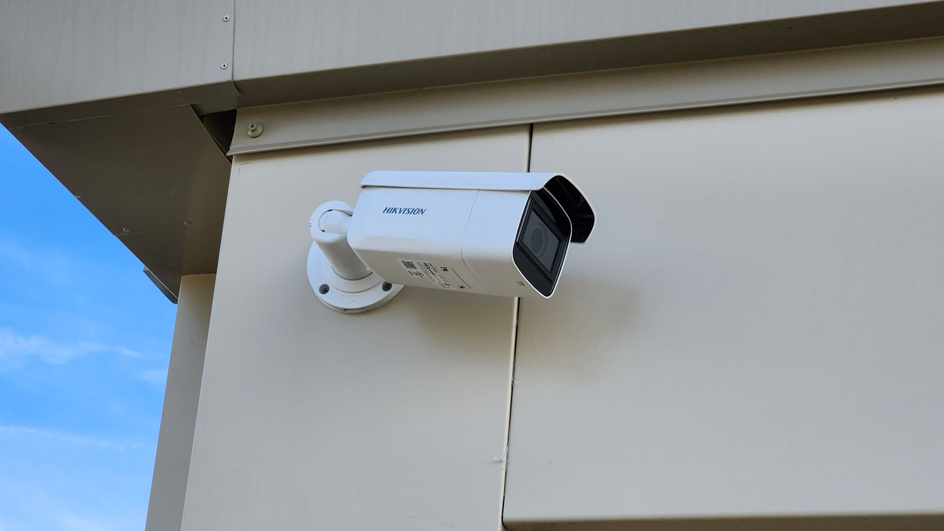White security camera mounted on a beige building exterior; blue sky visible.