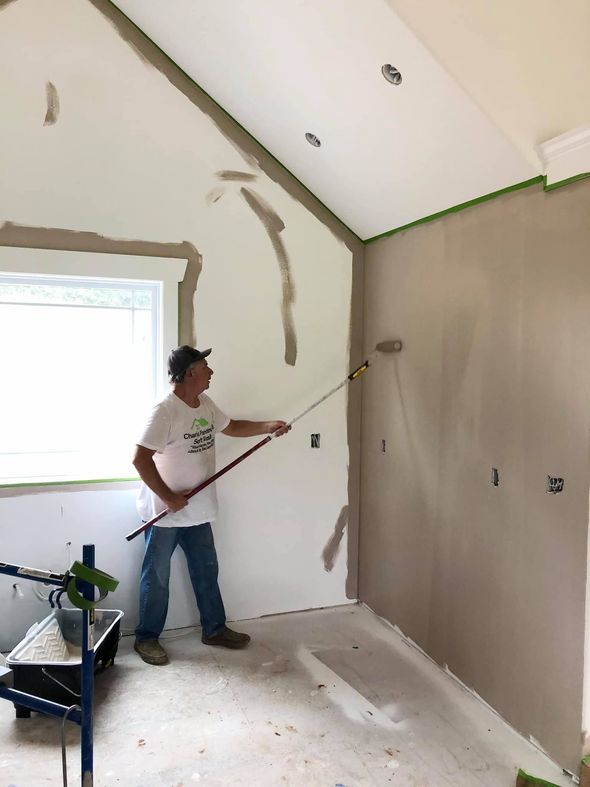Person painting a room with a roller, walls partly primed and protected with plastic.