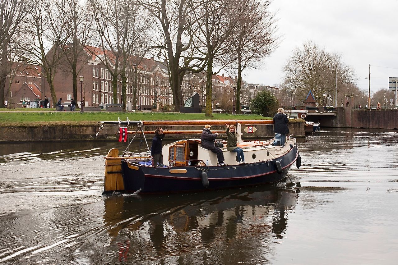 Een kanaalboot met passagiers vaart onder een brug door over een rustige rivier in een stadspark.