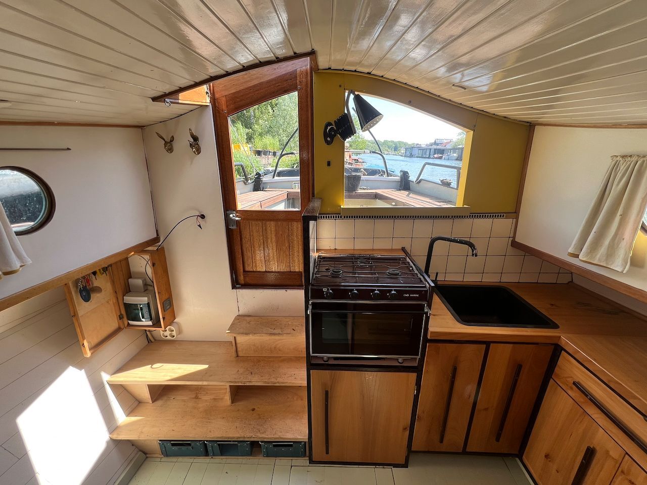 Compacte bootkajuit met houten kasten, fornuis, spoelbak en een lichte, openslaande deur naar het dek.