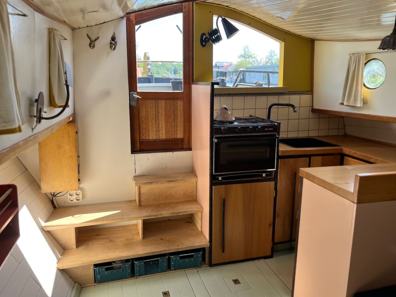 Kajuit met compacte keuken, houten banken en ramen met lichte gordijnen.