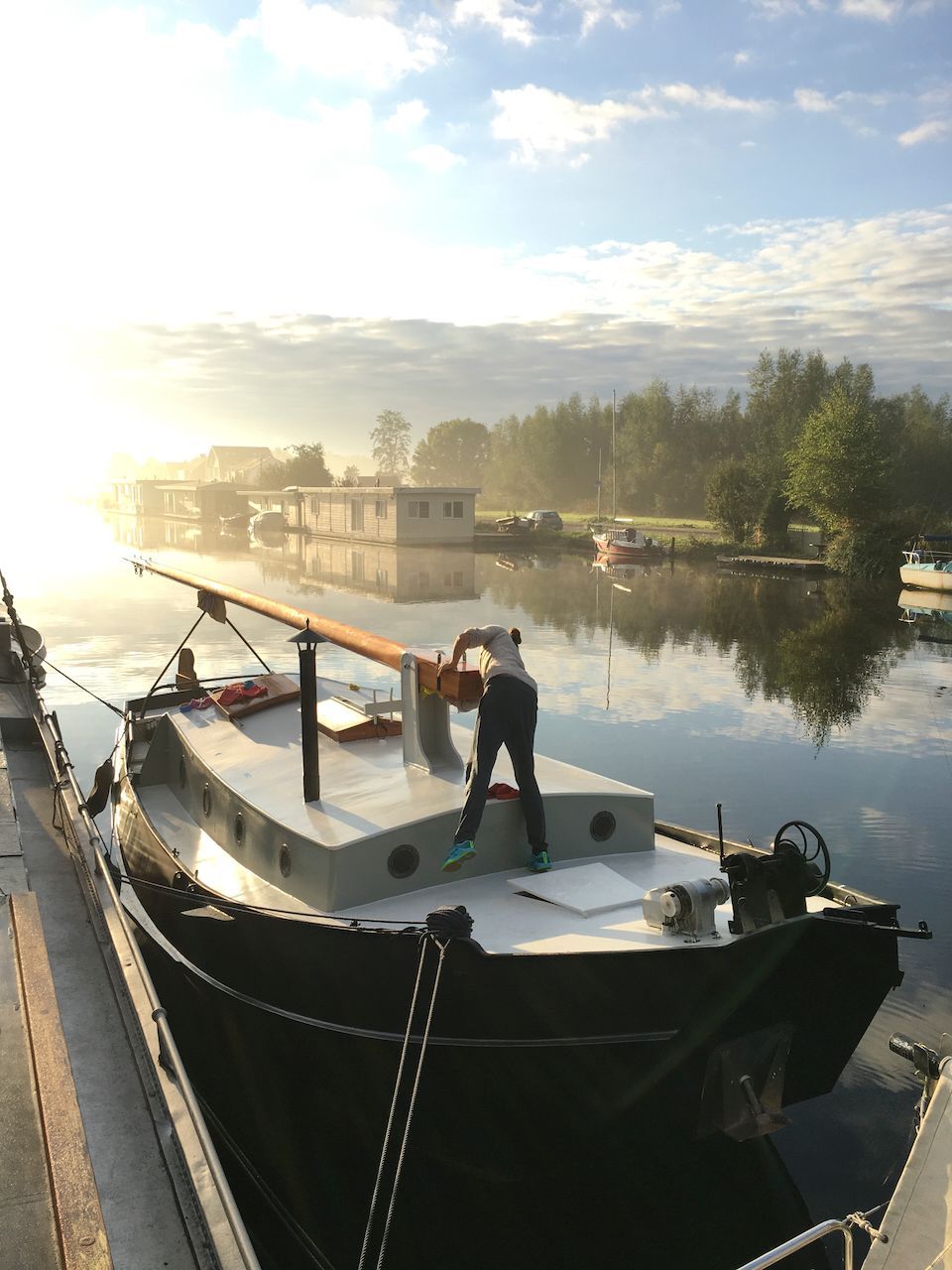 De Wijde Blik boot ligt aangemeerd op een kalme rivier bij zonsopgang, met een persoon die op het dek staat.