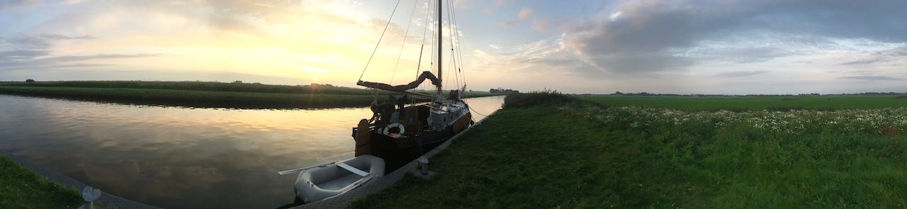 De Wijde Blik op kalm water bij zonsondergang, met een begroeide oever en een bewolkte hemel.