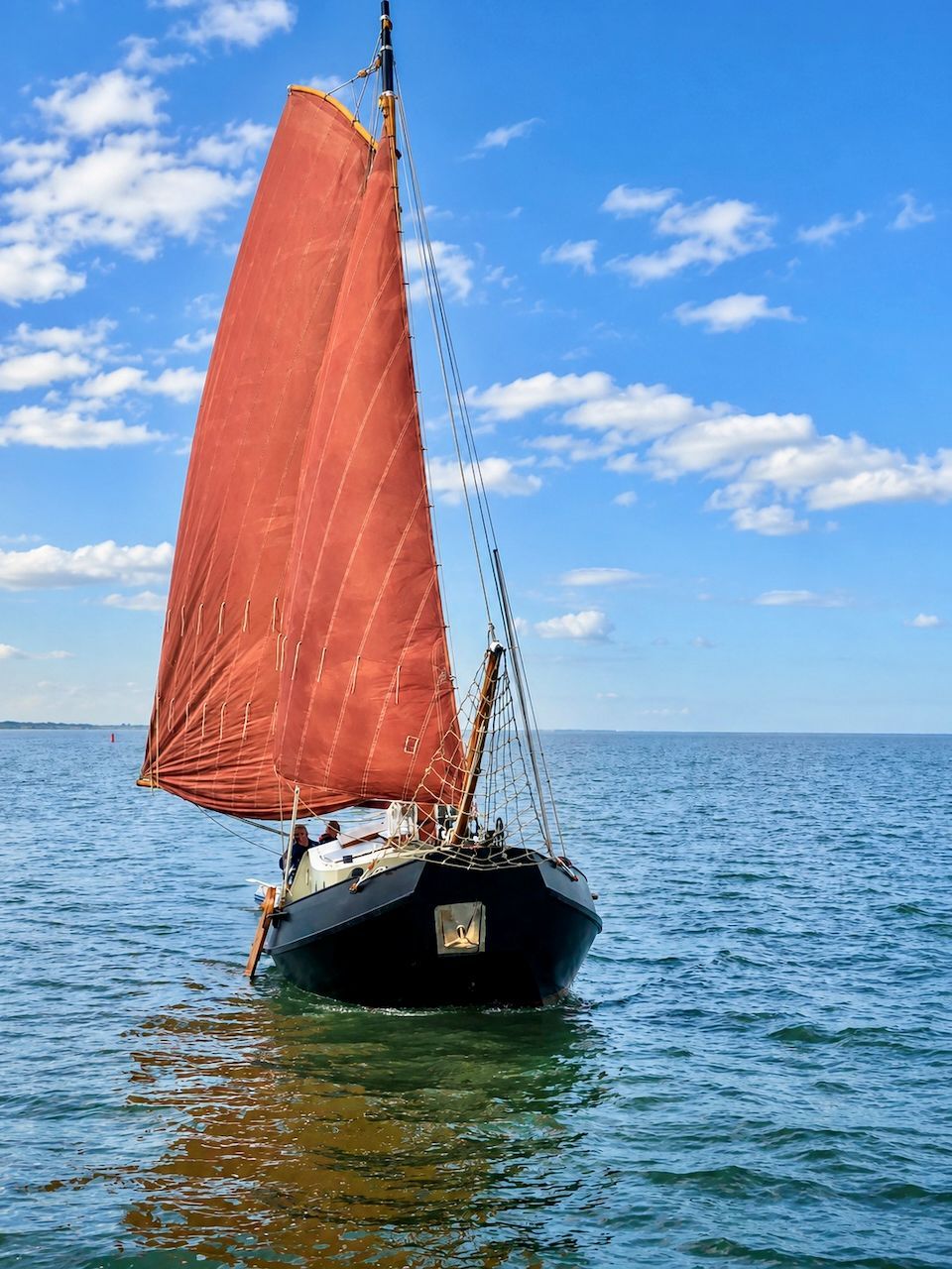 Een zeilboot met rode zeilen op kalm blauw water onder een gedeeltelijk bewolkte hemel.