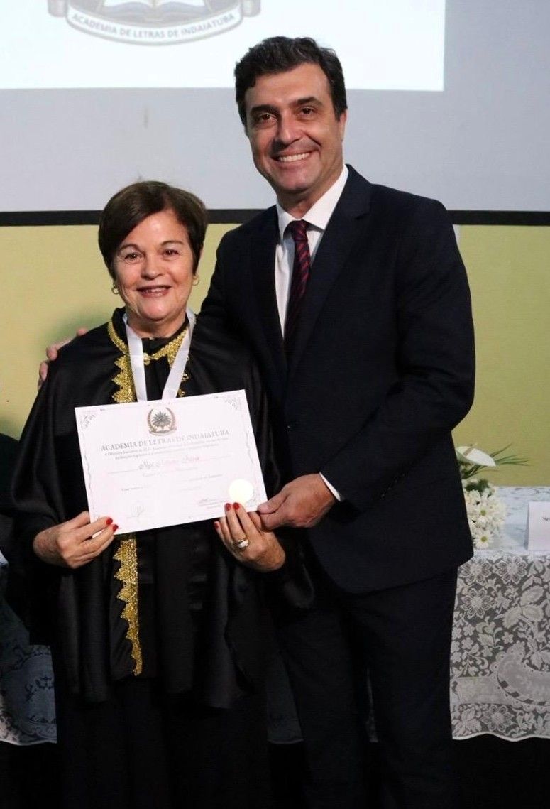 Mulher de beca de formatura segurando um certificado, sorrindo; homem de terno ao lado dela.
