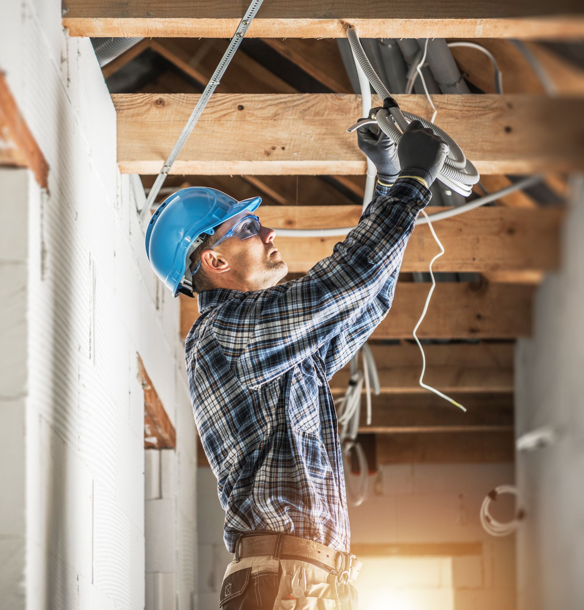 Construction worker in blue hard hat installing wiring in an unfinished ceiling.