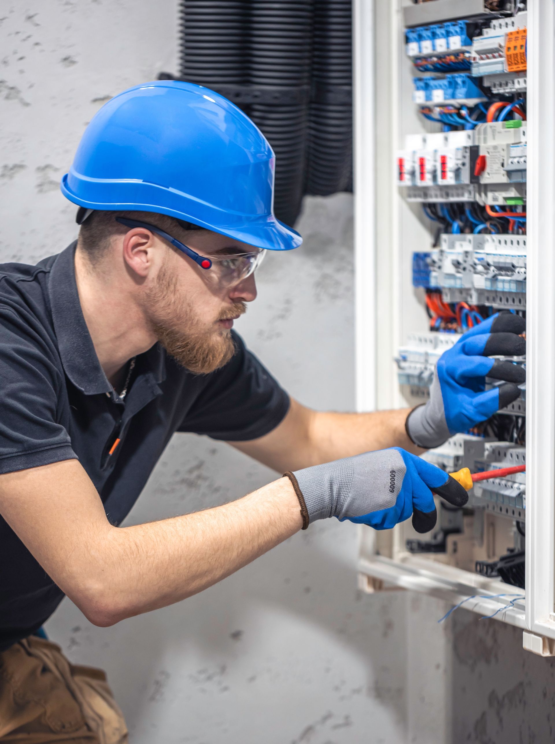 Electrician in blue hard hat and gloves wiring an open electrical panel.