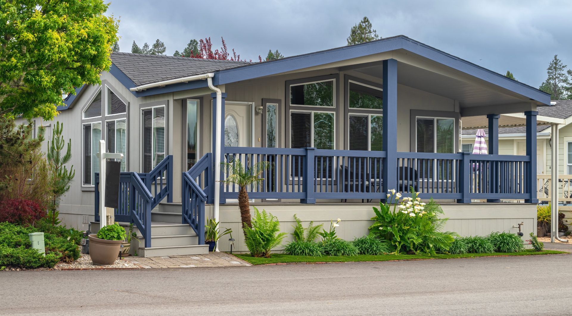 Blue-gray house with a wide porch, white trim, and lush green landscaping by a quiet street