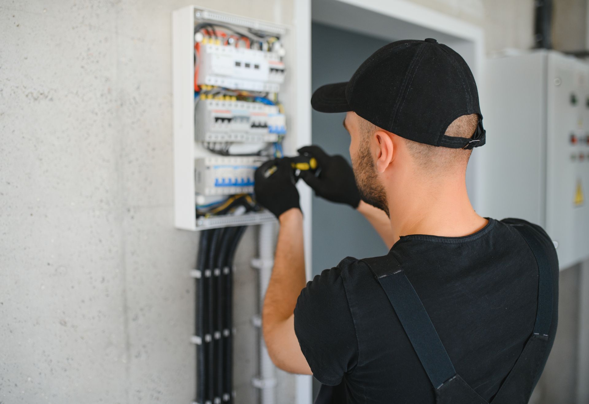 Electrician in black shirt and cap wiring an open electrical panel indoors