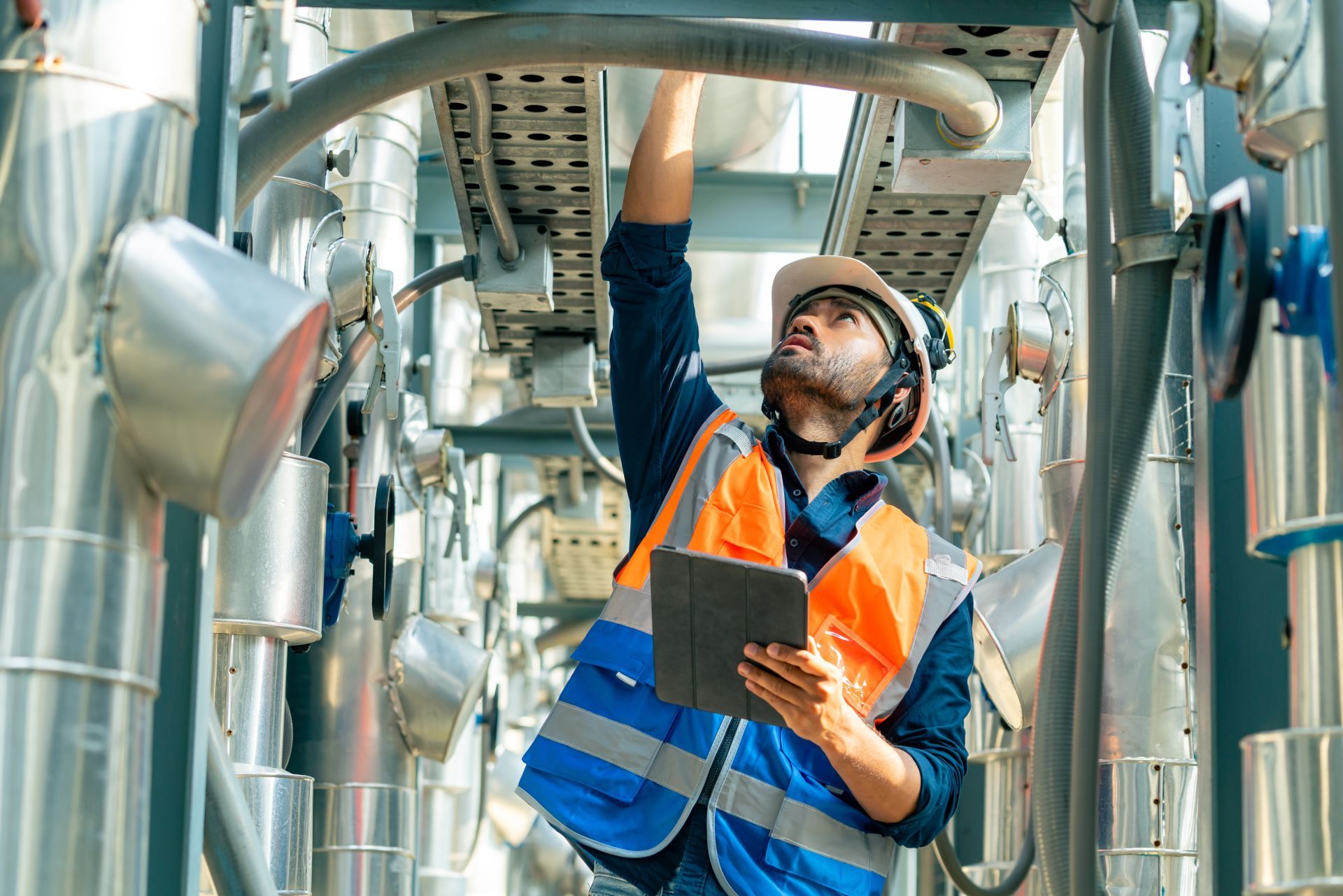 Worker in orange safety vest inspects overhead pipes in an industrial plant, holding a tablet.