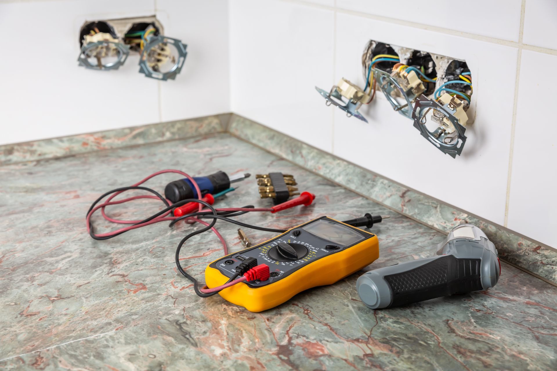 Tools on a countertop beside exposed electrical boxes and wiring in a kitchen wall