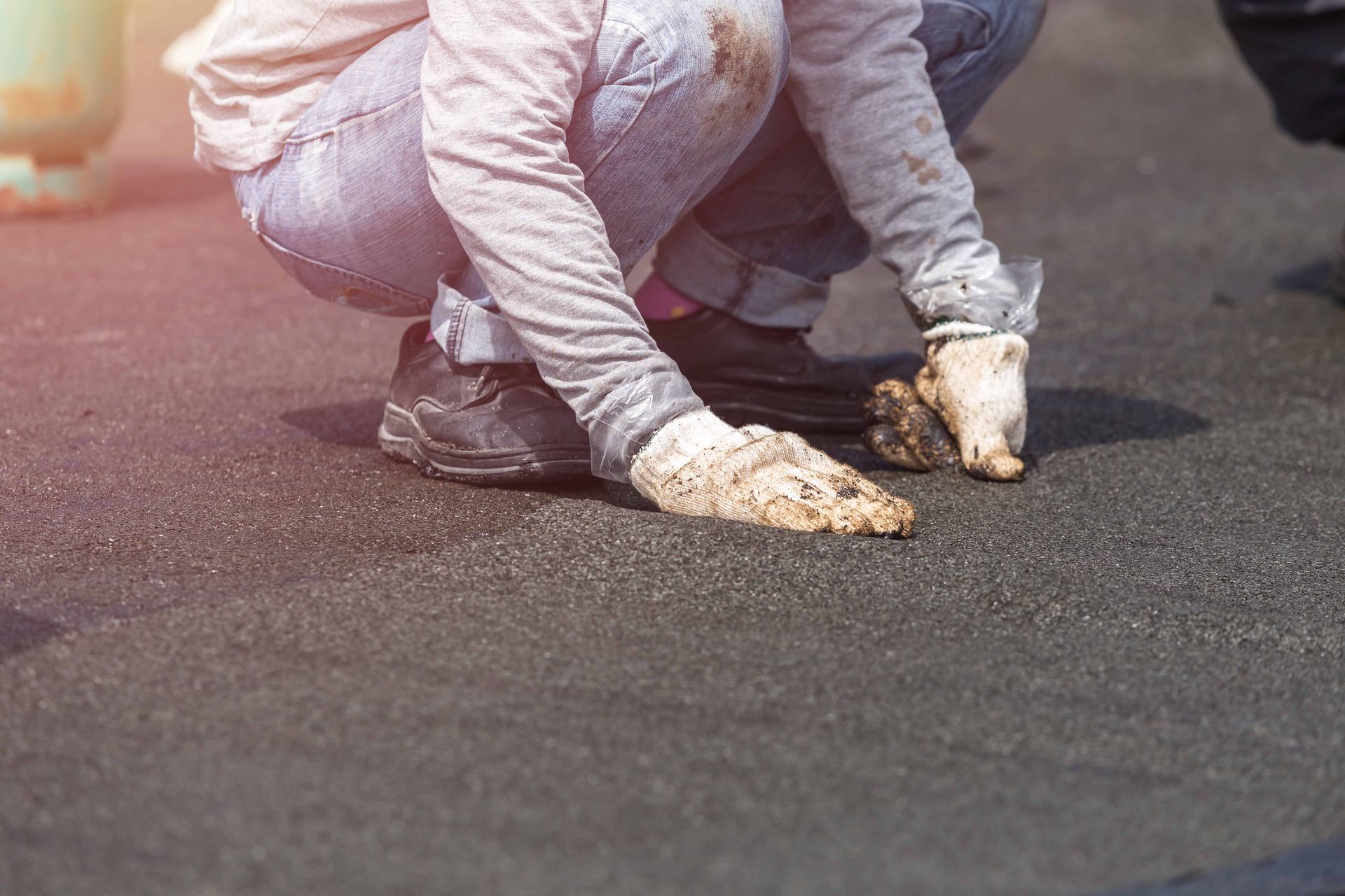 A person wearing gloves, kneeling on dark asphalt, possibly laying roofing.