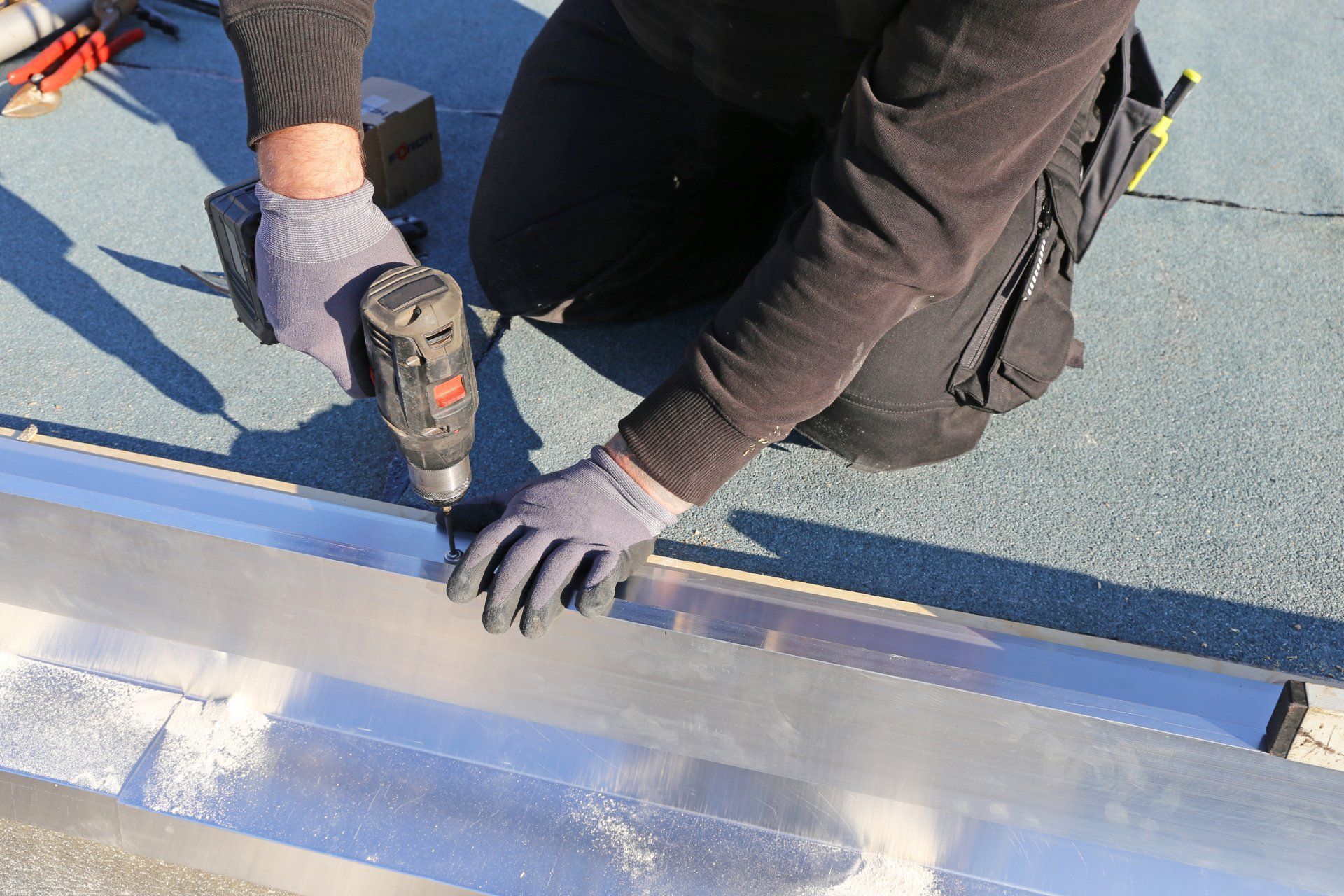 Person in work clothes using a drill to install metal flashing on a flat roof.