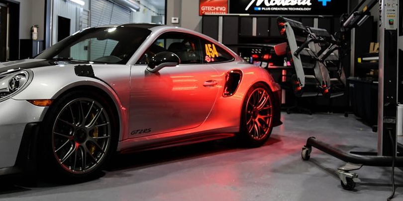 Silver sports coupe in a garage, lit by red underglow, with another car and tools in the background