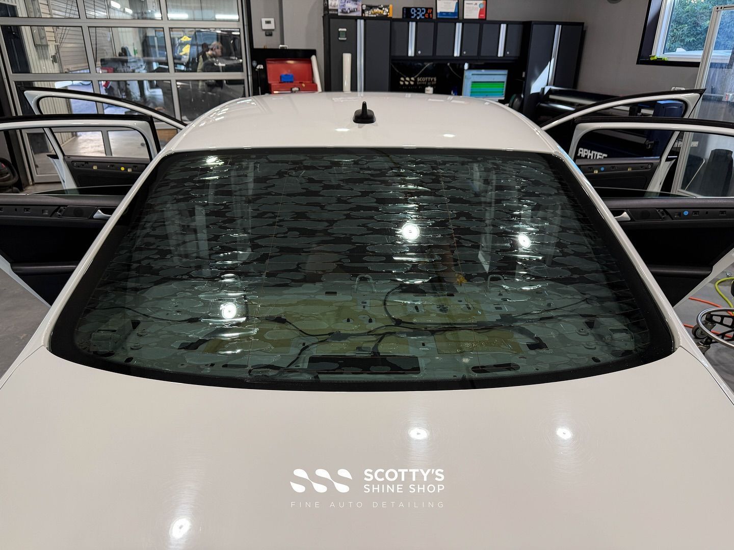 White car seen from the front in an auto shop, with a wet windshield and open doors in the background.