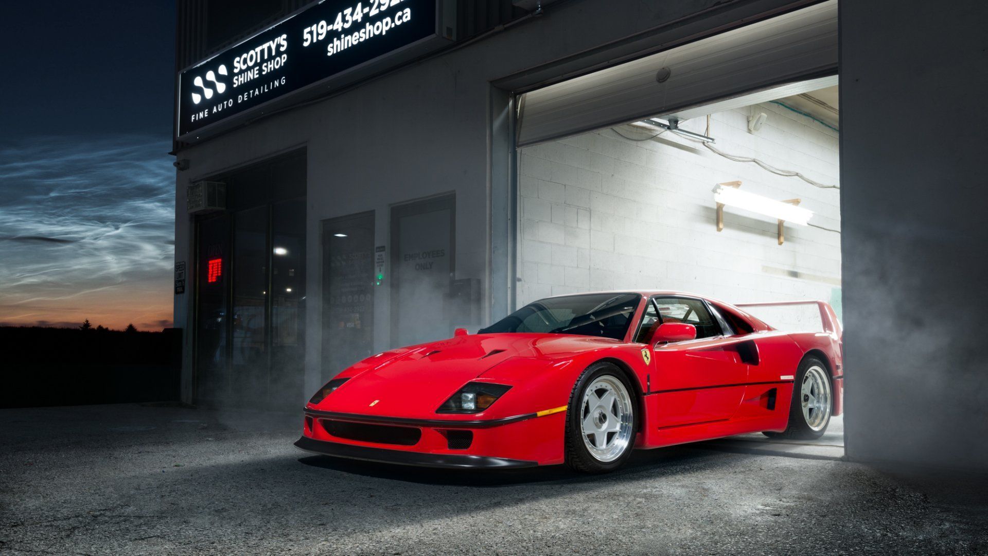Red sports car parked in a garage at dusk, with a glowing workshop interior behind it.
