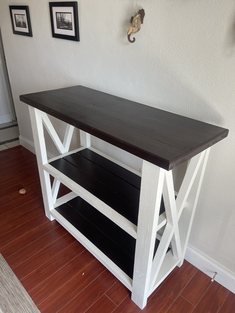 White X-frame console table with dark wood top and shelves against a wall on a hardwood floor
