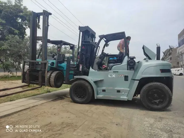 Una carretilla elevadora azul claro levanta una caja de madera al aire libre en una carretera pavimentada cerca de árboles y un edificio.