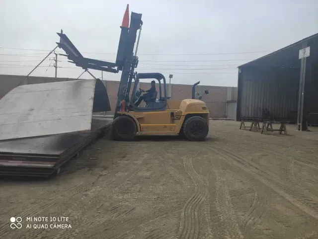 Una carretilla elevadora amarilla levanta un gran panel metálico en un patio industrial junto a un almacén.