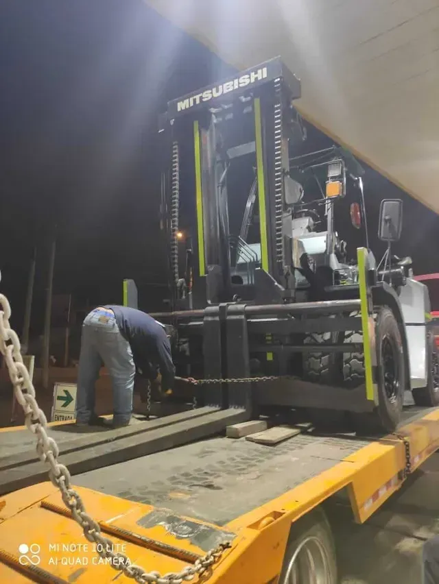 Un trabajador carga una carretilla elevadora Mitsubishi amarilla en un remolque de plataforma en un almacén o patio.