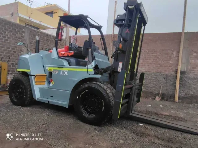 Una carretilla elevadora gris está estacionada junto a un muro de ladrillos en un aparcamiento al aire libre, con las horquillas levantadas en posición vertical.