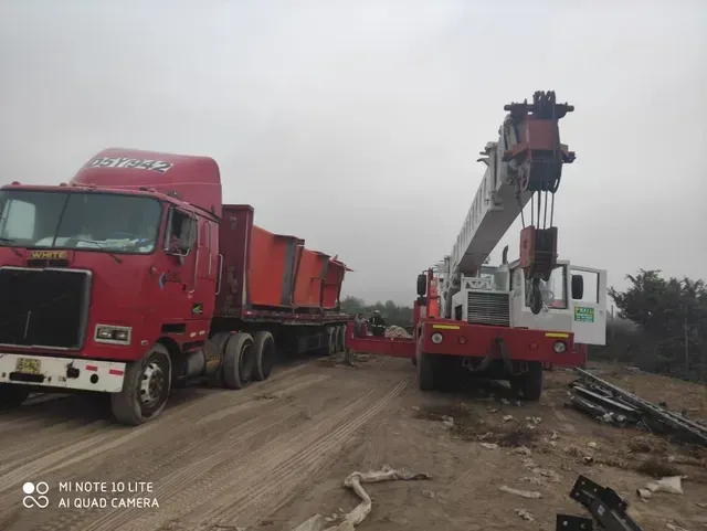 Camión volquete rojo junto a una grúa en un arcén embarrado bajo un cielo gris