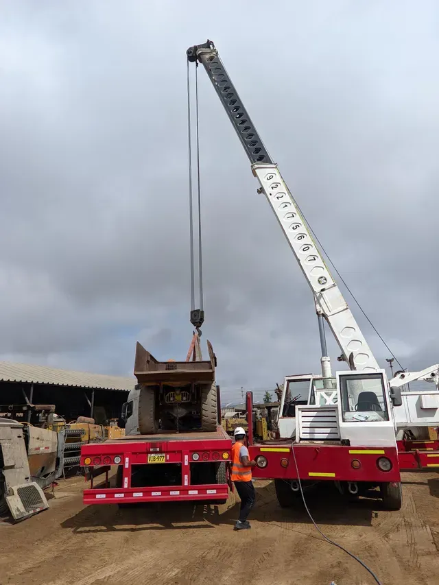 Una grúa está levantando una pequeña estructura de madera sobre un camión de plataforma roja en una obra, con trabajadores cerca.